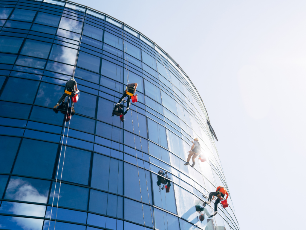 Window cleaners rappelling down the curved glass facade of a modern office building on a sunny day.