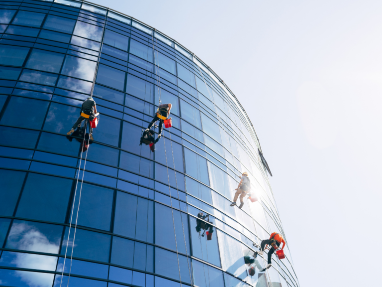 Window cleaners rappelling down the curved glass facade of a modern office building on a sunny day.